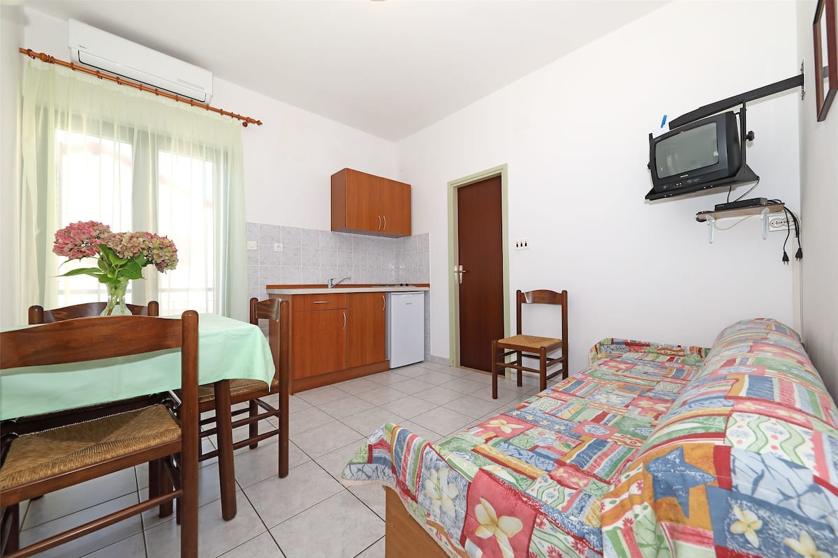 A compact room with a bed, a dining table with chairs, a small kitchenette, and a television mounted on the wall.