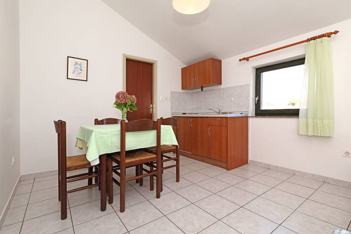 A compact kitchen with wooden cabinets, a dining table with chairs, and a window with a curtain.
