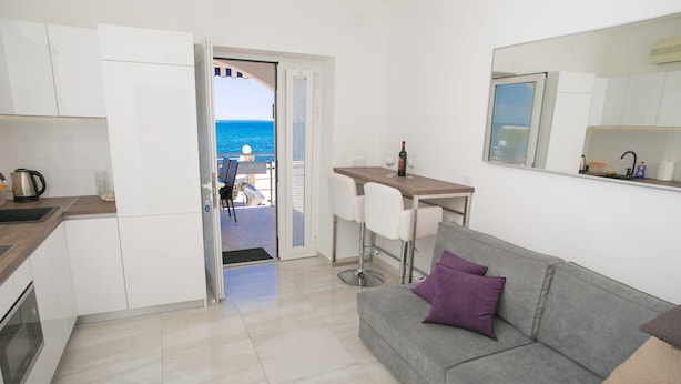 A compact living space with a kitchenette, a dining area, and a sofa with cushions. A door leads to an outdoor area with a view of the sea. - Apartment Primorska (Novalja)