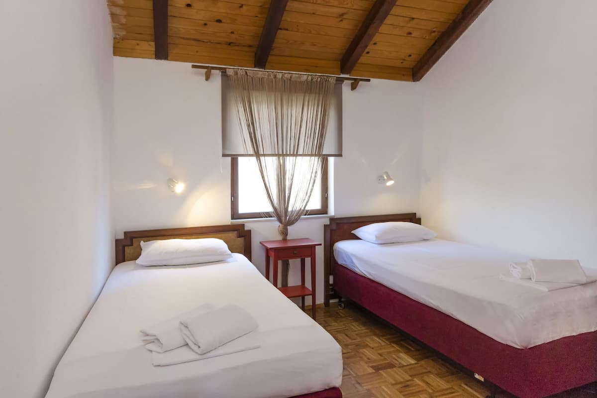 A room with two beds, wooden beams on the ceiling, a window with a curtain, and a small table with a vase.
