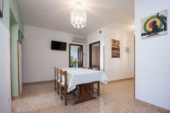 Comedor con mesa dispuesta para cuatro personas, una araña y un televisor fijado a la pared.