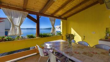 Un área de comedor al aire libre con techo, una mesa, sillas, una pared amarilla y vista al mar.