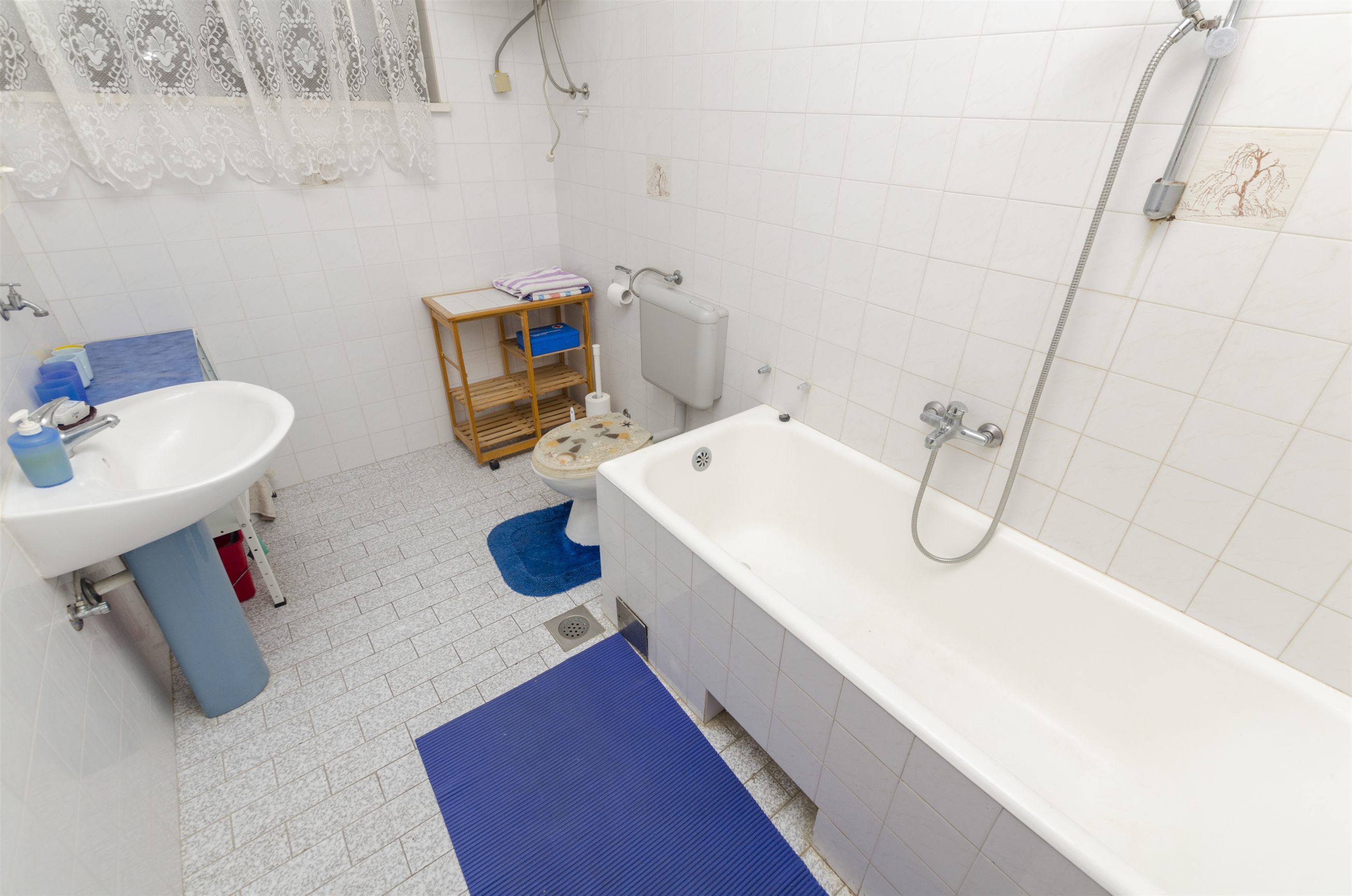 A bathroom with a white sink, a bathtub, a shower, and a toilet. There is a blue mat on the floor and a wooden shelf with toiletries.