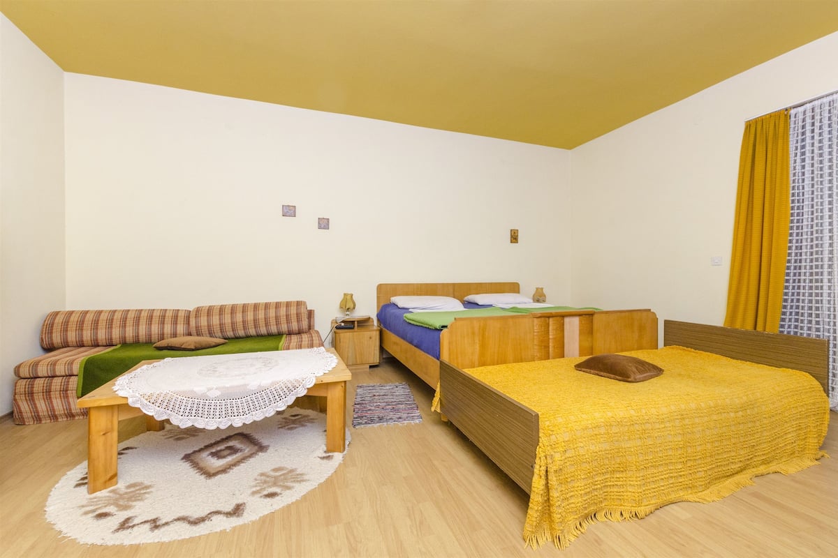 A bedroom with a sofa, a bed with a yellow blanket, a wooden nightstand, and a small table with a lace tablecloth.