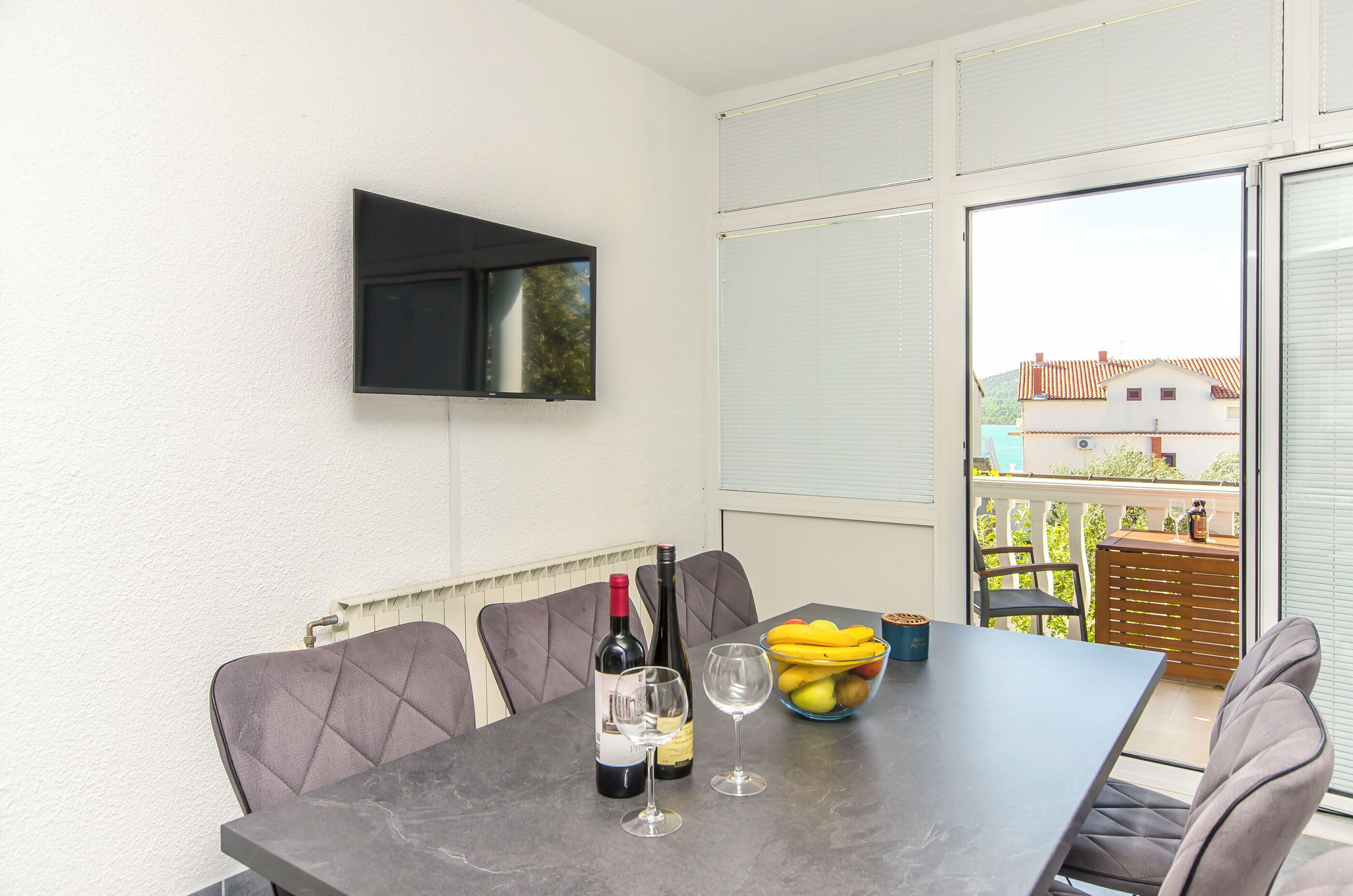 A balcony with a table, chairs, a bottle of wine, a bowl of fruit, and a TV mounted on the wall.