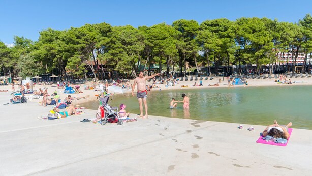 Una playa con gente tomando el sol y nadando, rodeada de árboles y edificios al fondo.