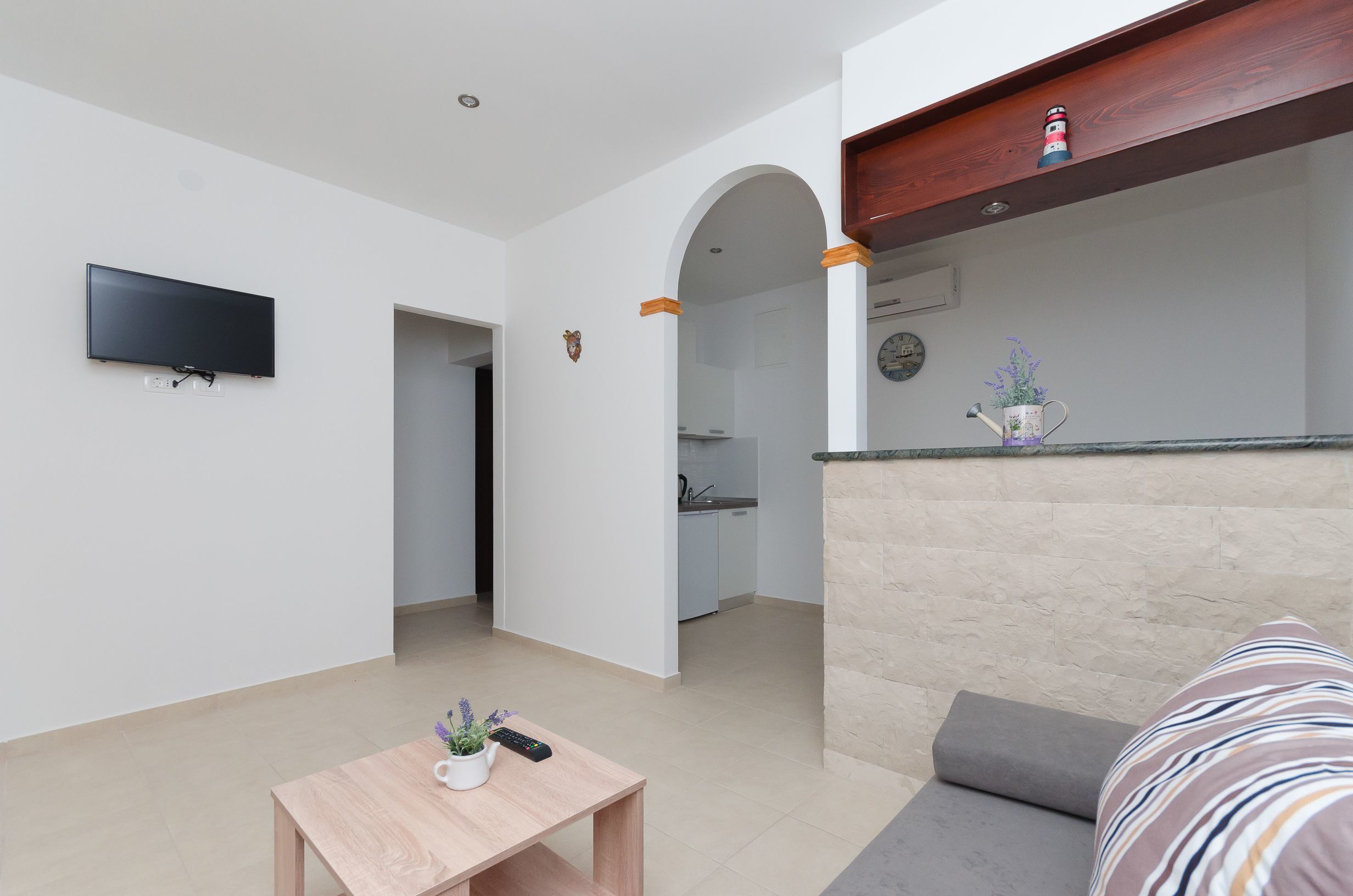 A modern living room with a flat-screen TV mounted on the wall, a wooden coffee table with a remote and a small plant, a grey sofa with striped cushions, and an arched doorway leading to a kitchen area.