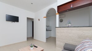 Ein modernes Wohnzimmer mit einem an der Wand befestigten Flachbildfernseher, einem Couchtisch aus Holz mit Fernbedienung und einer kleinen Pflanze, einem grauen Sofa mit gestreiften Kissen und einer Bogenöffnung zu einer Küche.