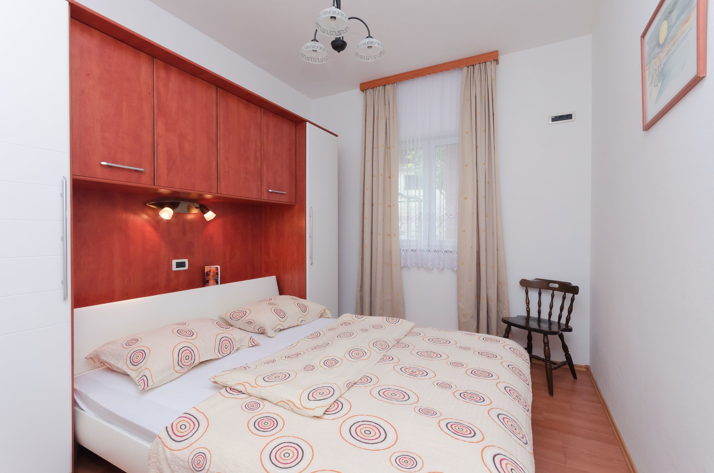 Ein Schlafzimmer mit einem Bett, einem Holzschrank, einem Sessel und einem Fenster mit Vorhängen.