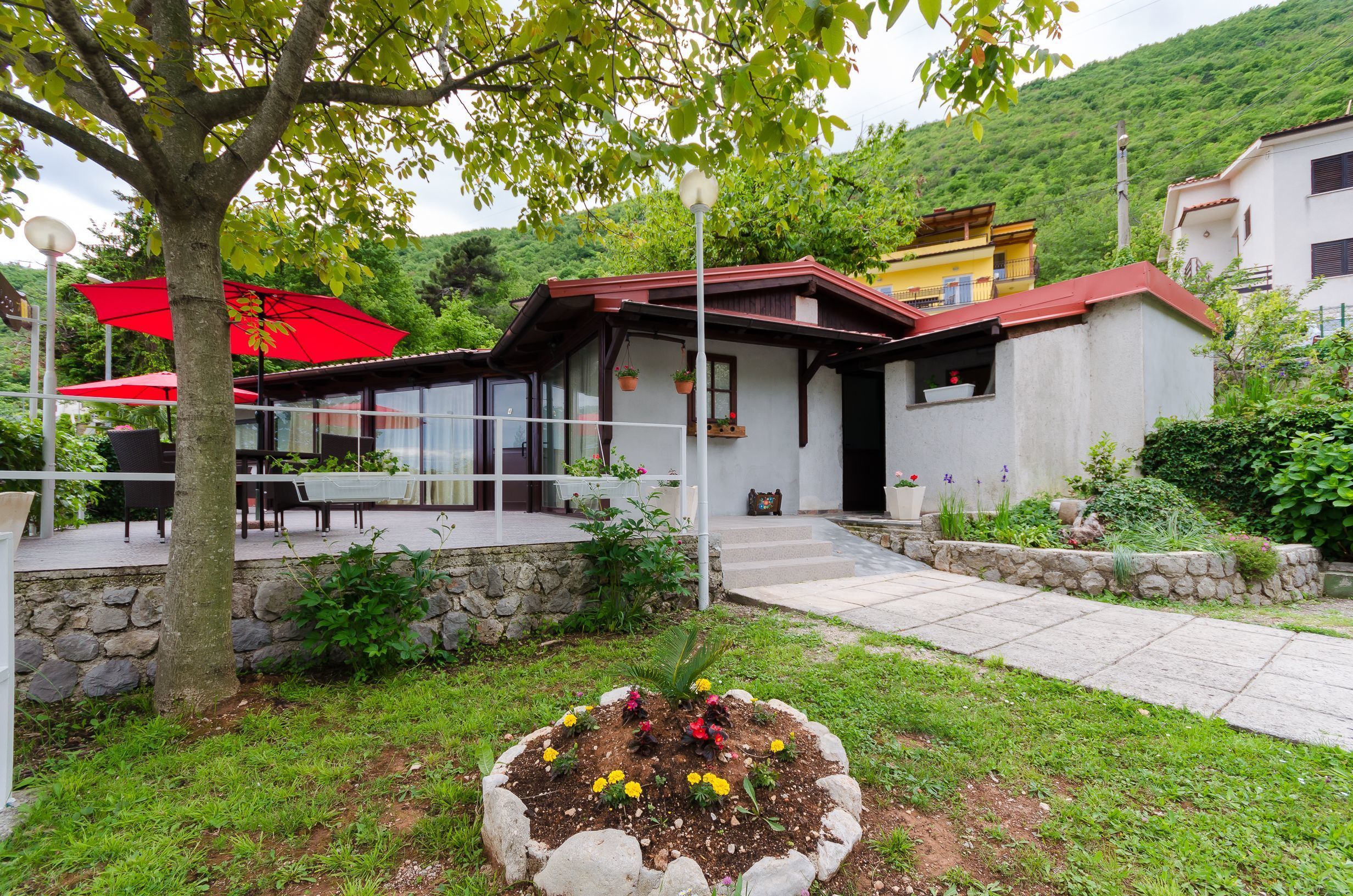 A house with a stone wall, a garden with flowers, and a red umbrella.