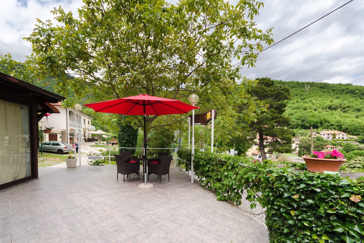 Eine Terrasse im Freien mit einem roten Sonnenschirm, zwei Stühlen und einem Blumentopf.