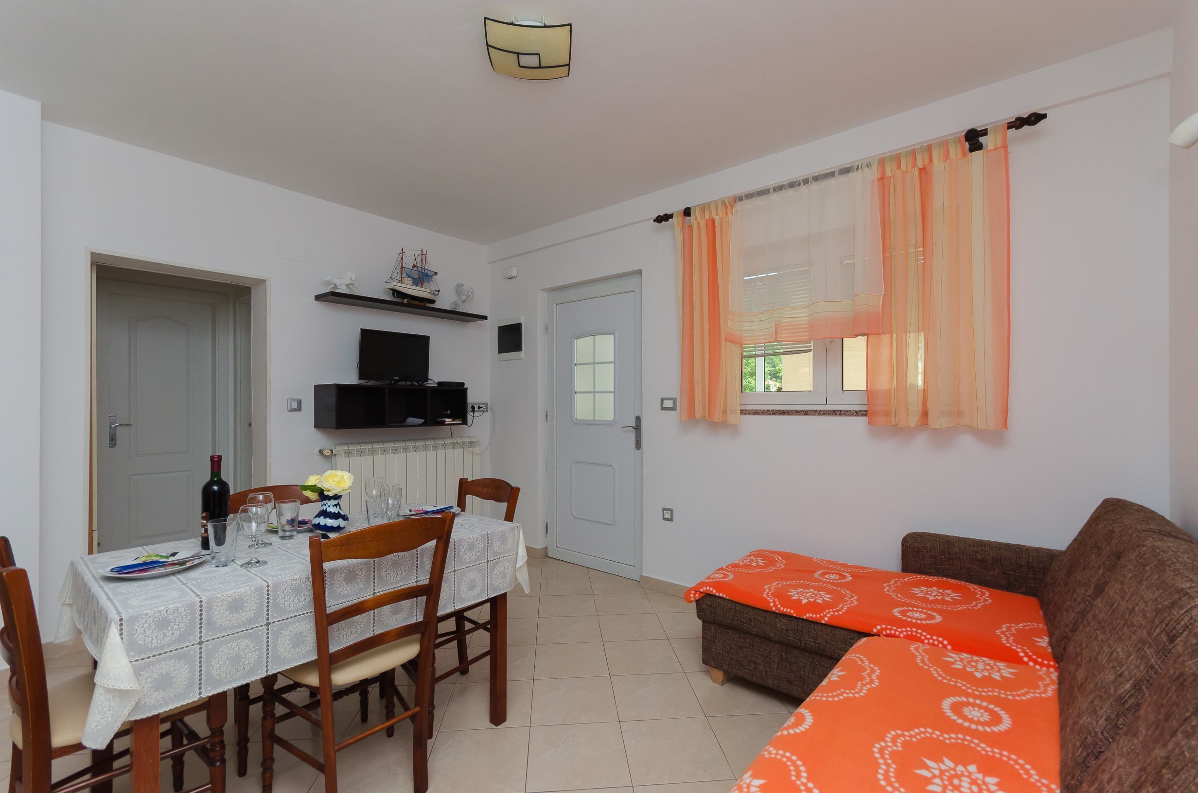 A dining area with a table set for two, a television above the table, and a window with orange curtains.