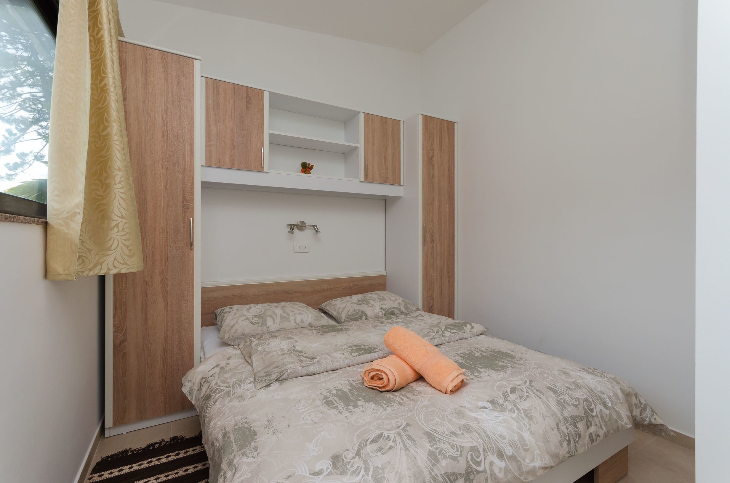 A bedroom with a bed, a wooden headboard, a small shelf, and a window with curtains.