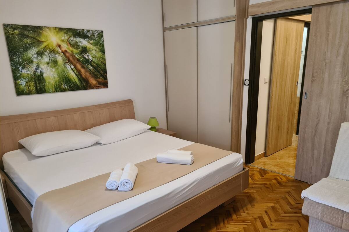 Un lit bien fait, avec des draps blancs et deux serviettes enroulées, une tête de lit en bois, une lampe de chevet verte et un tableau représentant une forêt accroché au mur.
