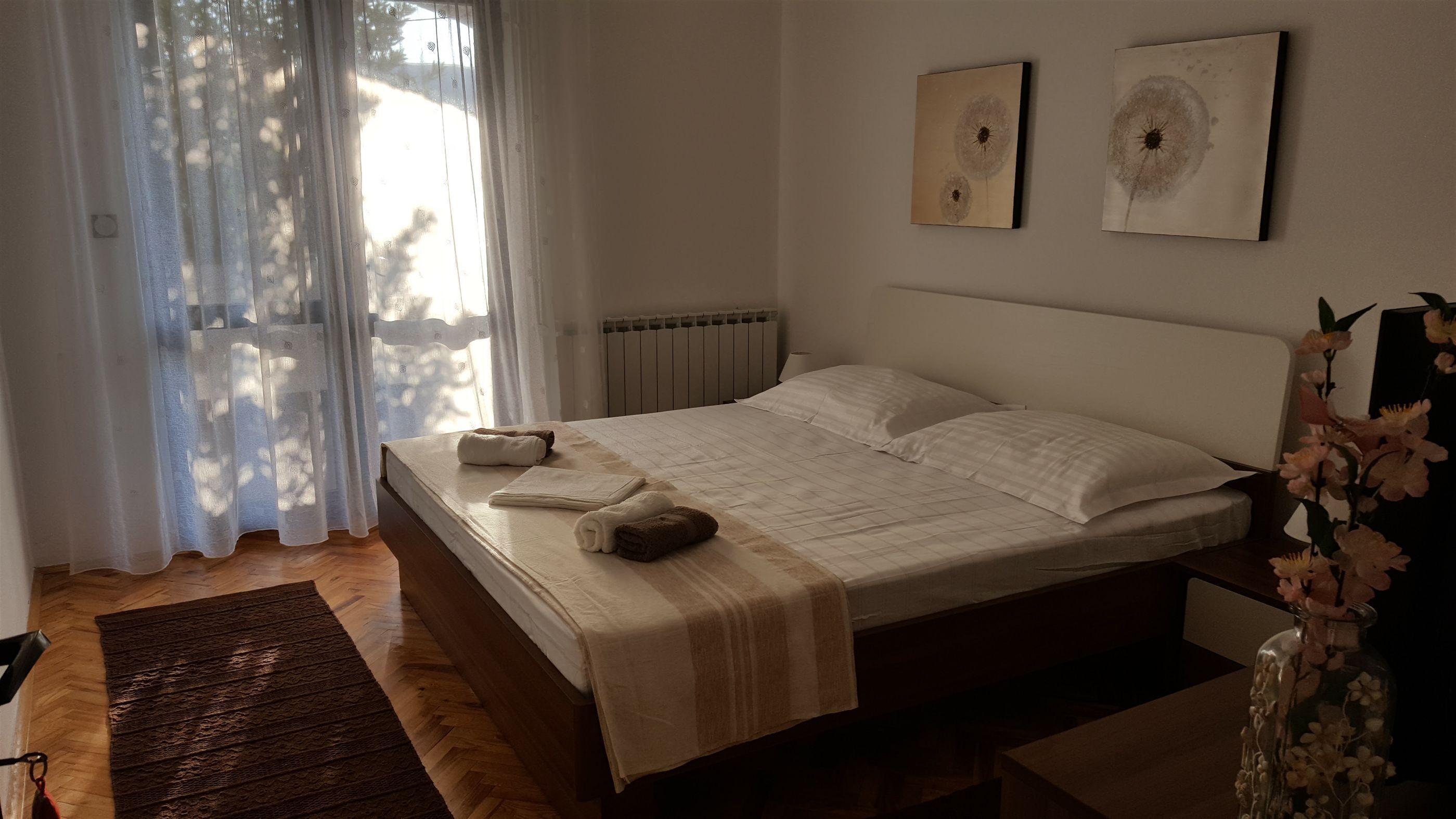 A bedroom with a bed, a radiator, a window with sheer curtains, and two wall-mounted artworks.