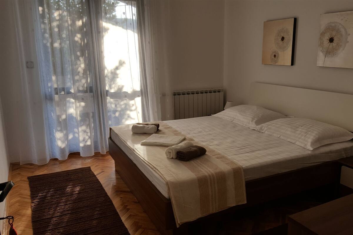 A bedroom with a bed, a radiator, a window with sheer curtains, and two wall-mounted artworks.