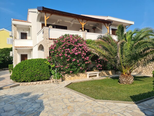 Una casa de dos pisos con fachada de piedra, un balcón y un jardín con una palmera y plantas con flores.