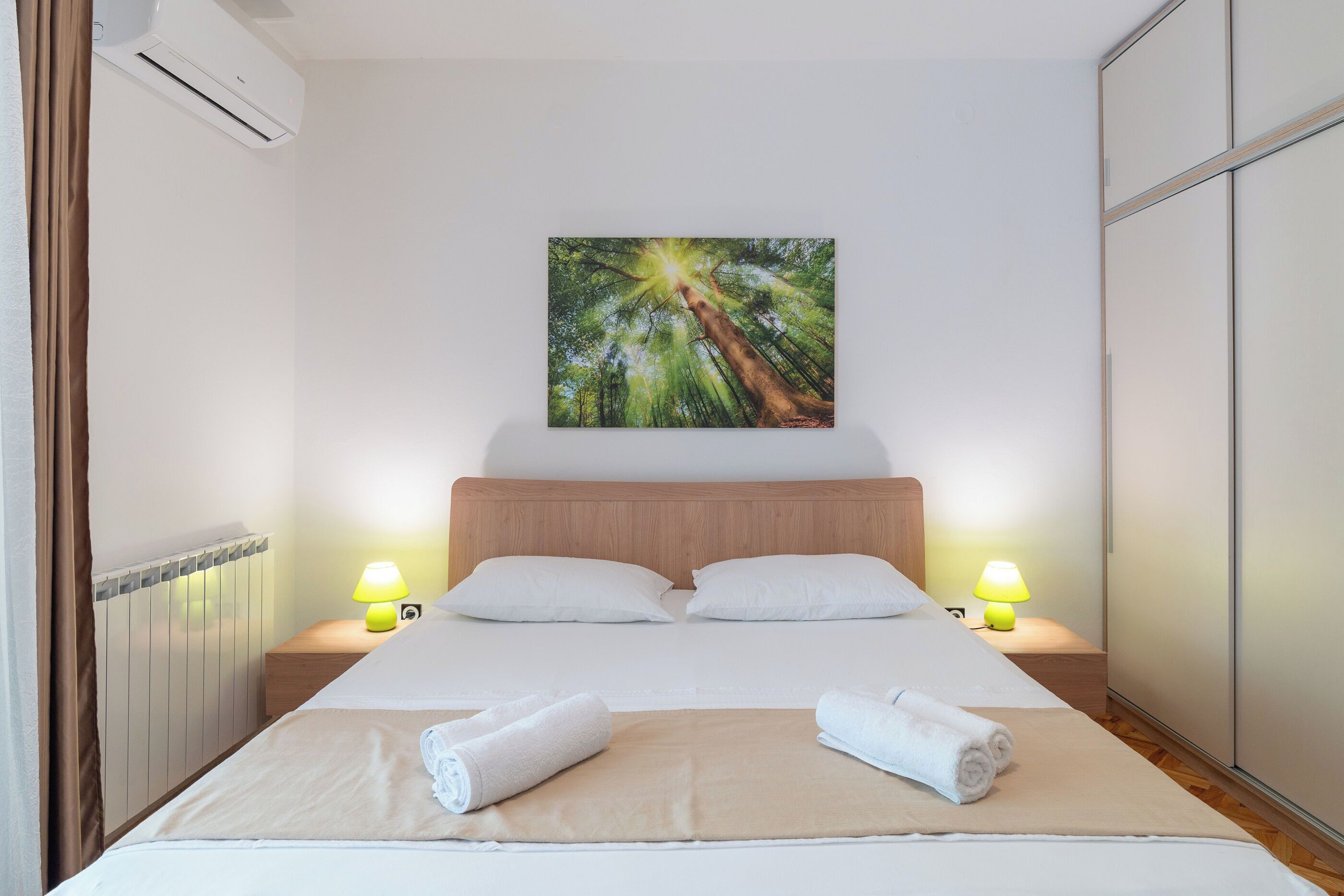 A hotel room with a bed, two bedside lamps, a painting on the wall, and two rolled towels on the bed.