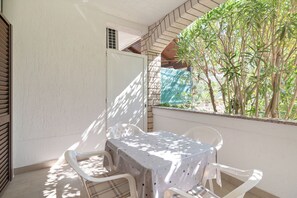 Un balcón con mesa y sillas, vistas a árboles y una ventana con cortina.
