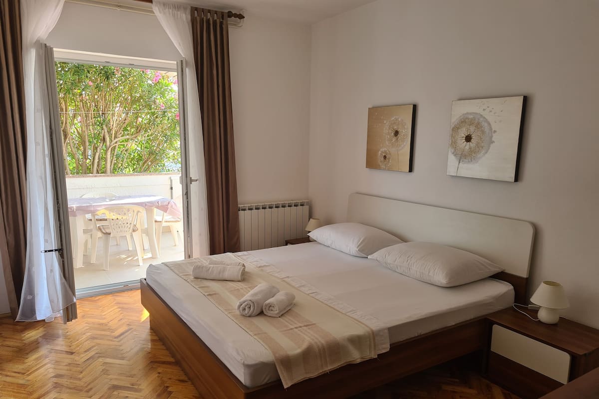 Un lit bien fait, avec du linge de lit blanc et deux oreillers, une table de chevet avec une lampe, et une vue sur une terrasse extérieure avec des chaises et une table.