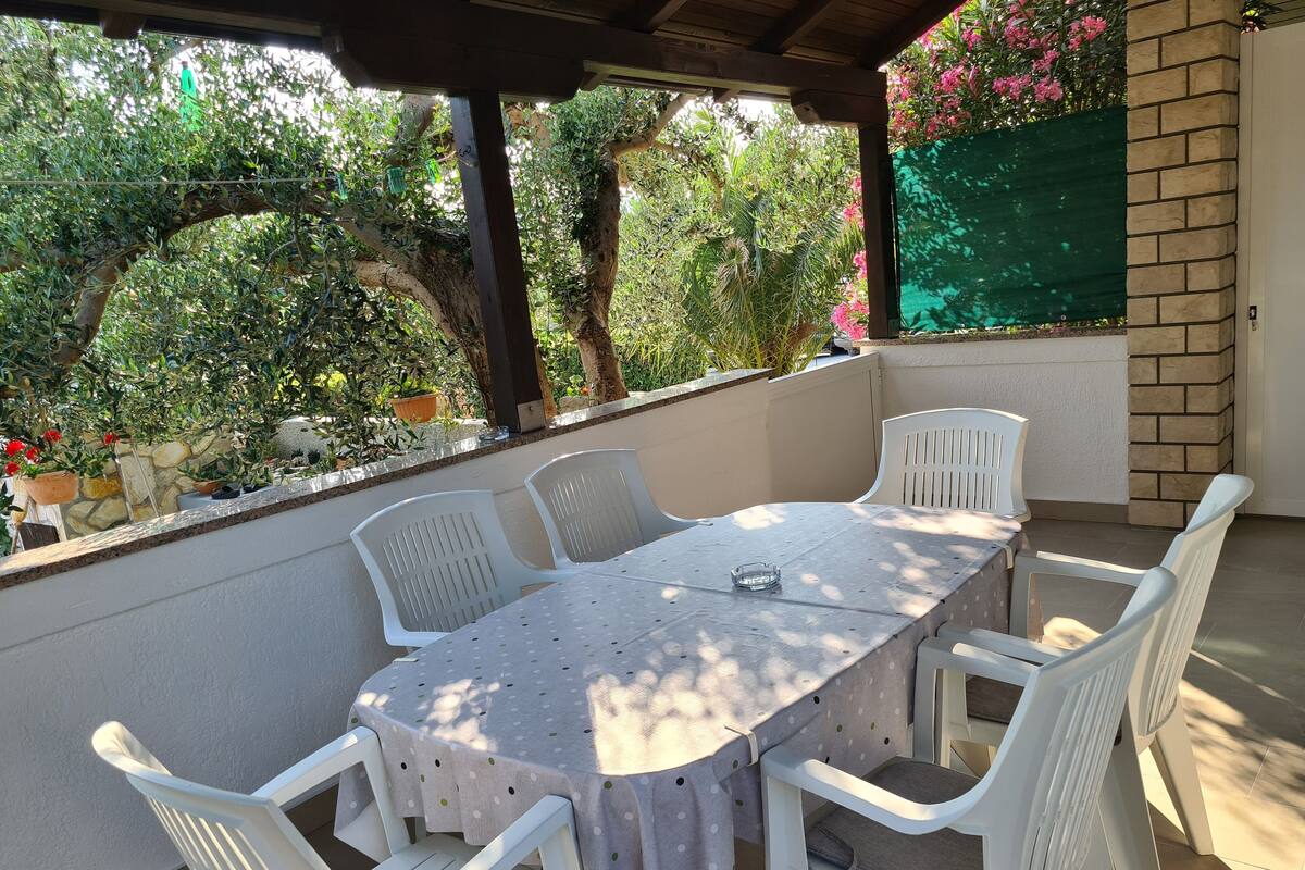 Une terrasse extérieure couverte, avec une table ronde et des chaises en plastique blanches, entourée de plantes en pot et d’un mur en briques.