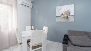 Ein Zimmer mit Esstisch, Stühlen, einem grauen Sofa, einer Klimaanlage, einem Bild von einem Boot und einer Fernbedienung auf dem Sofa.