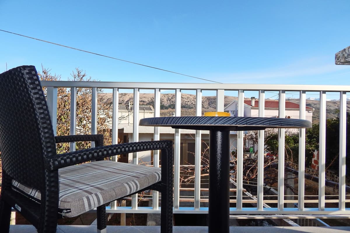 A balcony with a wicker chair and a small table, overlooking a residential area with houses and hills.