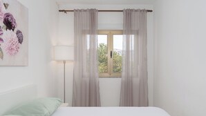 Ein Schlafzimmer mit einem Bett, einem Fenster mit Vorhängen, einer Lampe und einem floralen Bild an der Wand.