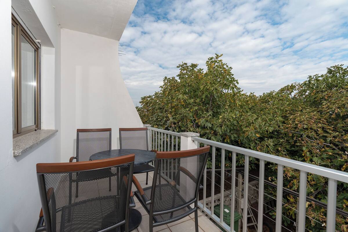 A balcony with a table and chairs, a white wall, and a view of trees.