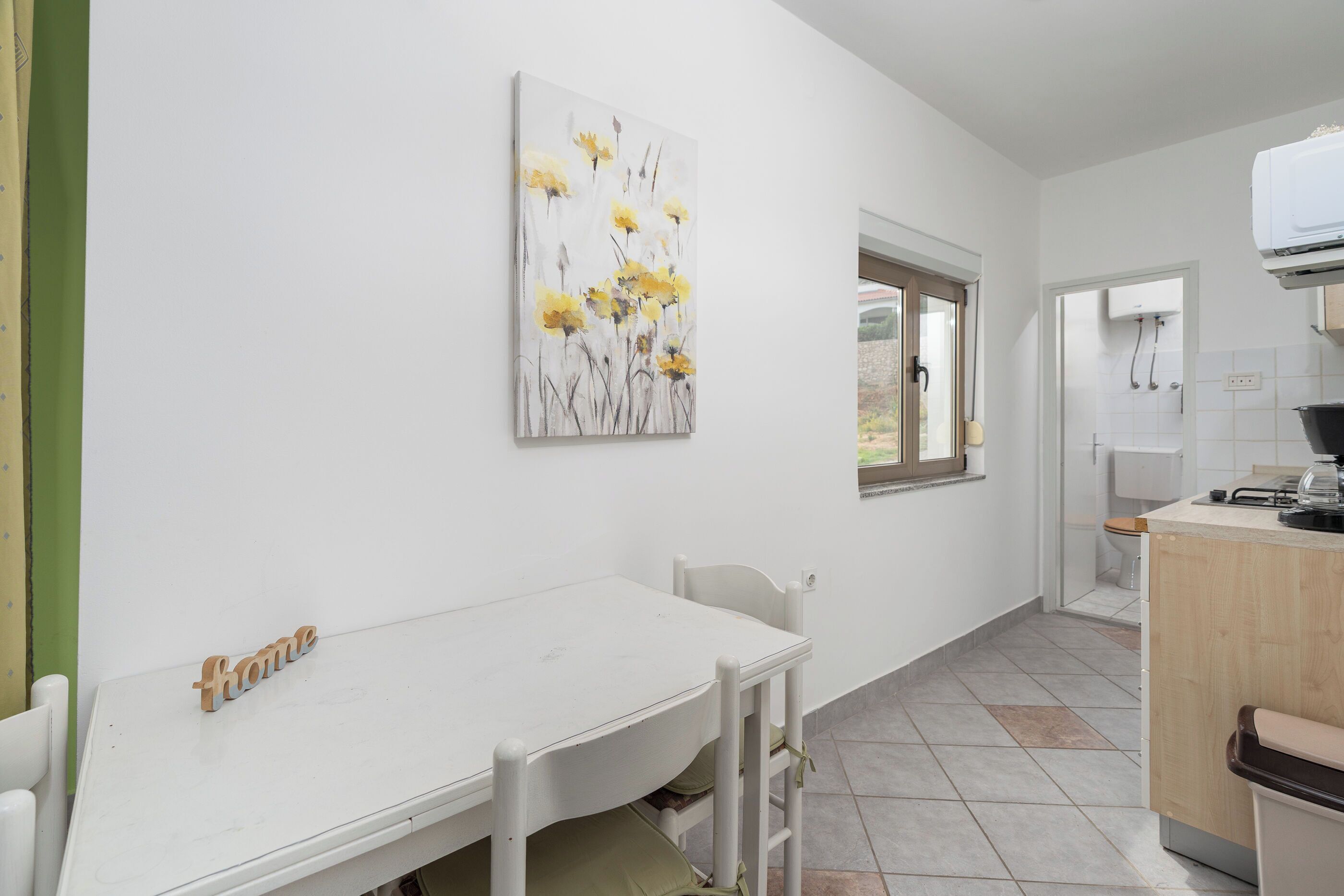Una mesa de comedor blanca con un letrero que dice 'home', un cuadro de flores amarillas y una cocina con un lavabo y encimera.