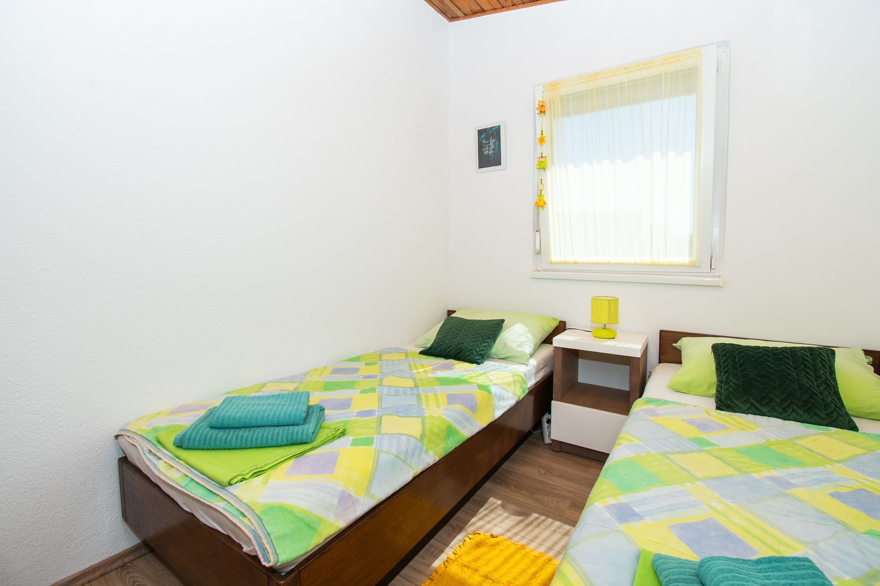A small, clean room with two single beds, a nightstand with a lamp, and a window with sheer curtains.
