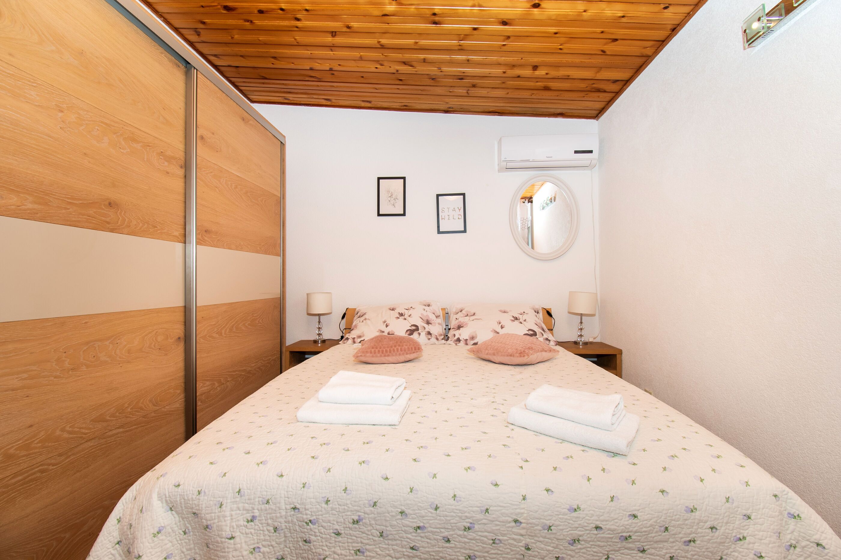 A bedroom with a wooden ceiling, a bed with floral bedding, two bedside lamps, and a wall-mounted air conditioner.