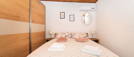 A bedroom with a wooden ceiling, a bed with floral bedding, two bedside lamps, and a wall-mounted air conditioner.
