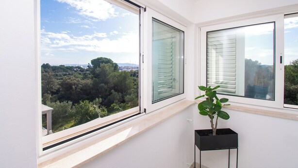 A room with large windows offering a view of a forested area and a distant bridge. A potted plant is placed on a small black stand near the windows. - Apartment Danijela (Bibinje)