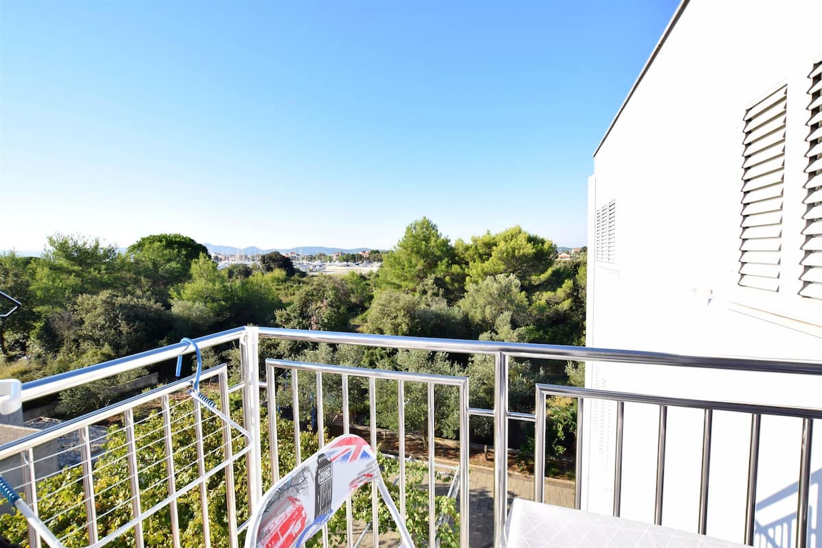 Ein Balkon mit Metallgeländer, ein weißes Gebäude mit Fensterläden und Blick auf eine grüne Landschaft mit Bäumen und entfernten Gebäuden.