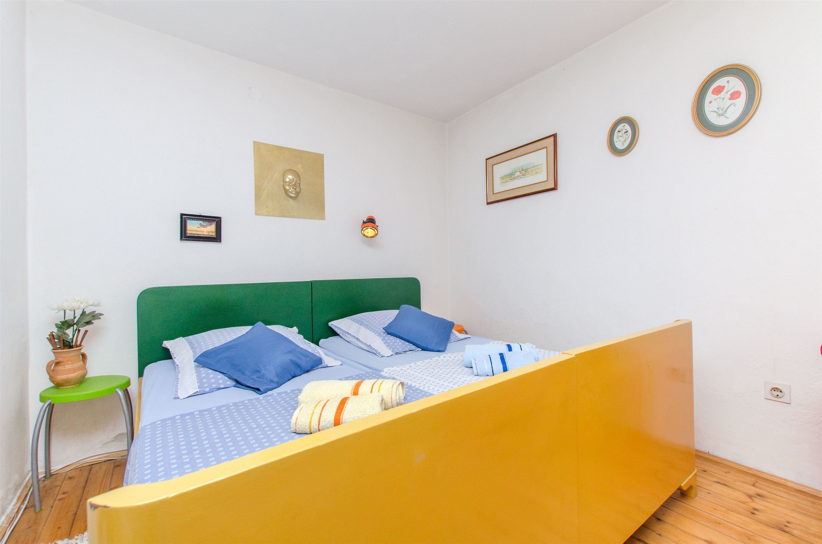 A room with a bed, a green stool, and a small table with a plant.