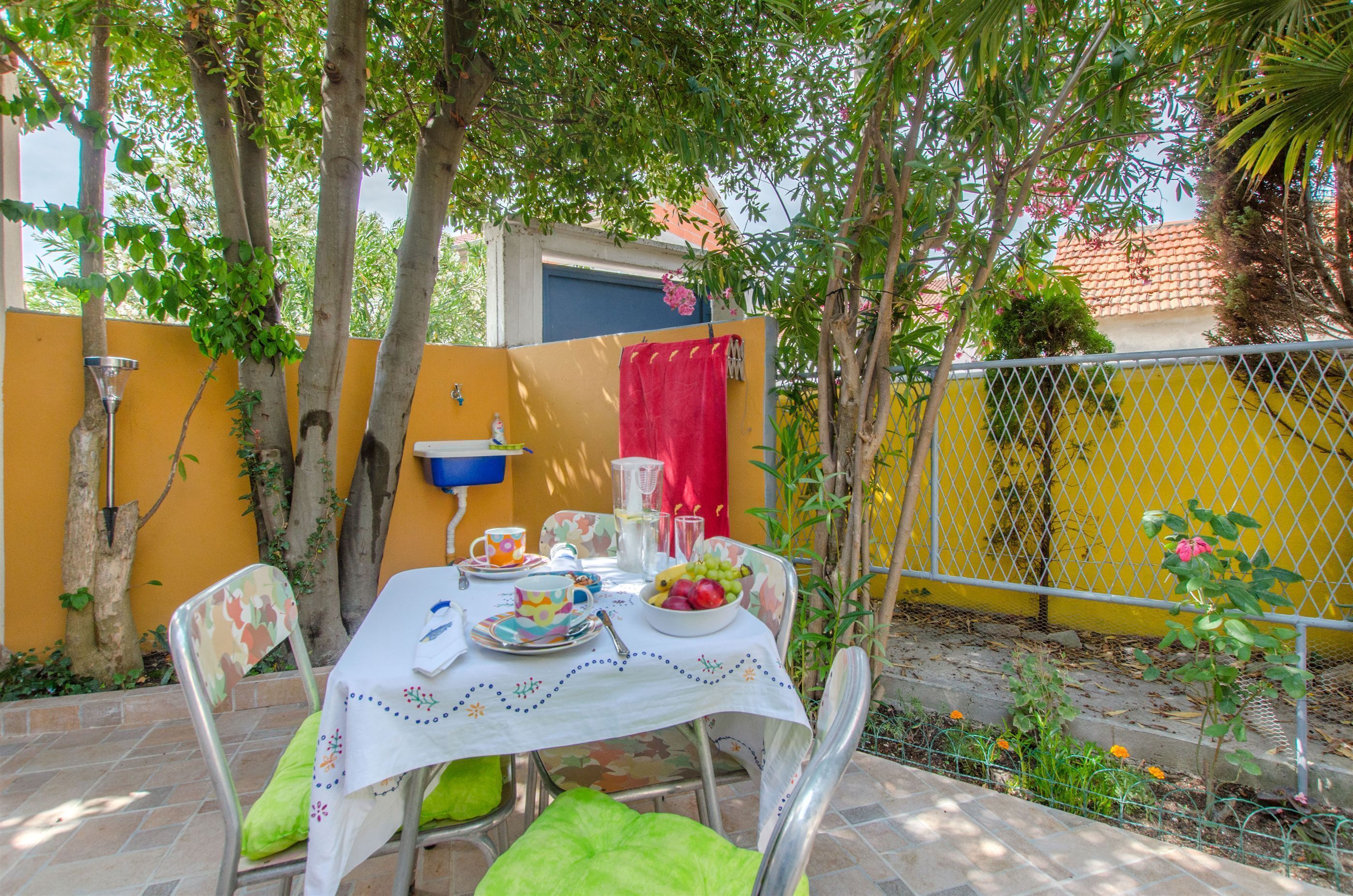 A patio with a table set for two, surrounded by chairs, a yellow wall, and a metal fence.