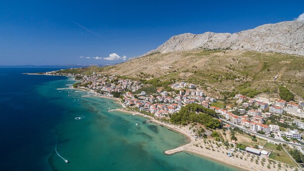An aerial view of a coastal town with buildings along the shoreline, a clear blue sea, and a mountainous backdrop. - Apartments Amulic (Duce)