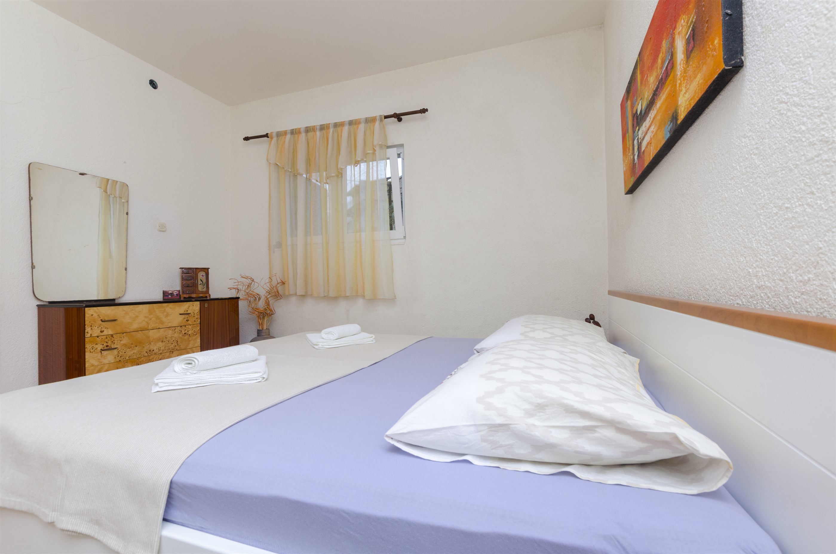 A bedroom with a bed, a dresser, a mirror, a window with curtains, and a painting on the wall.