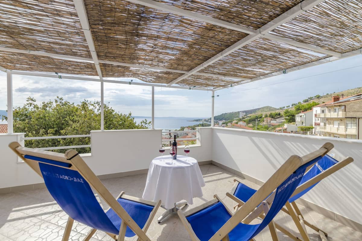A balcony with a table set for two, two blue chairs, and a view of the sea and hills.