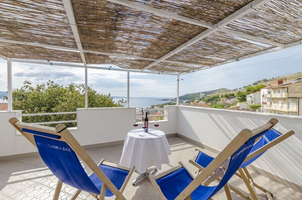 A balcony with a table set for two, two blue chairs, and a view of the sea and hills.