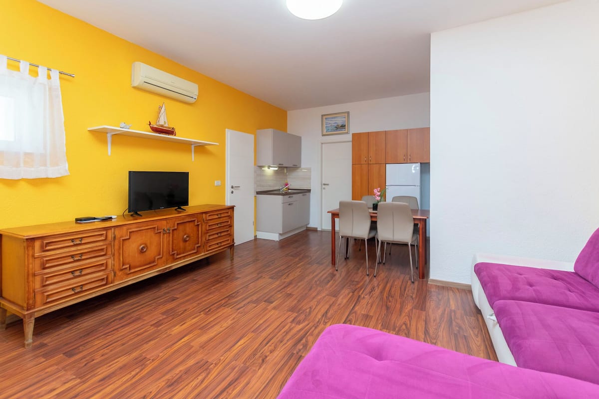 A room with a wooden cabinet, a television, a dining table with chairs, a purple sofa, and a kitchen area with cabinets and an oven.