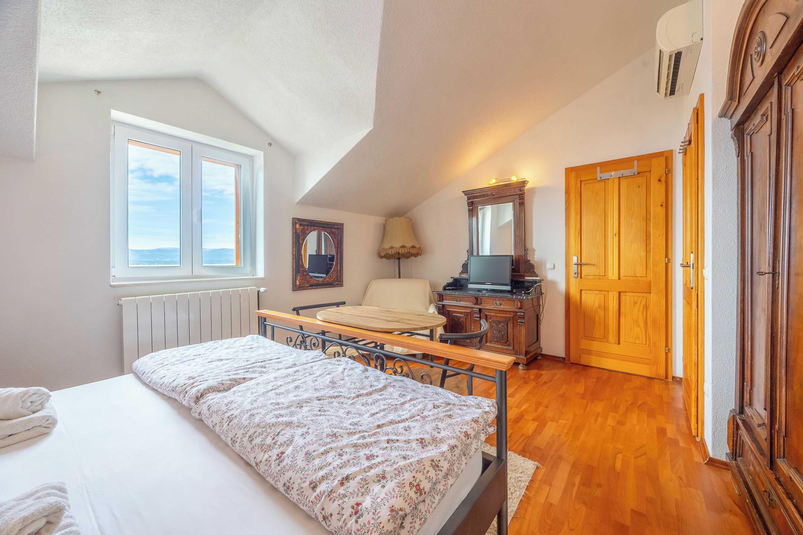 A bedroom with a bed, a desk, a chair, a wooden wardrobe, and a window.