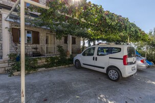 Un coche blanco estacionado en una entrada de casa, con un edificio de piedra y un árbol de hojas verdes al fondo.