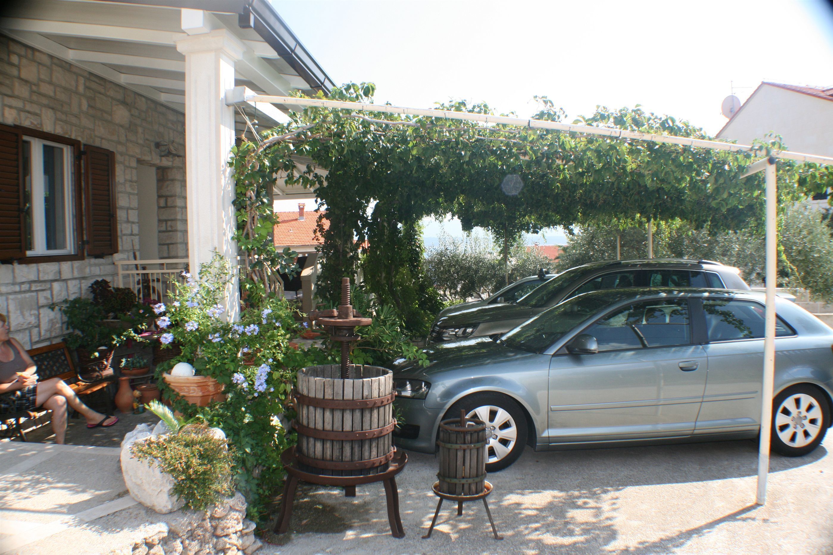 Ein Auto parkt auf einer Auffahrt mit einer von Grünwerk überwucherten Pergola, einem hölzernen Fass-Pflanzgefäß und einem Blumentopf.