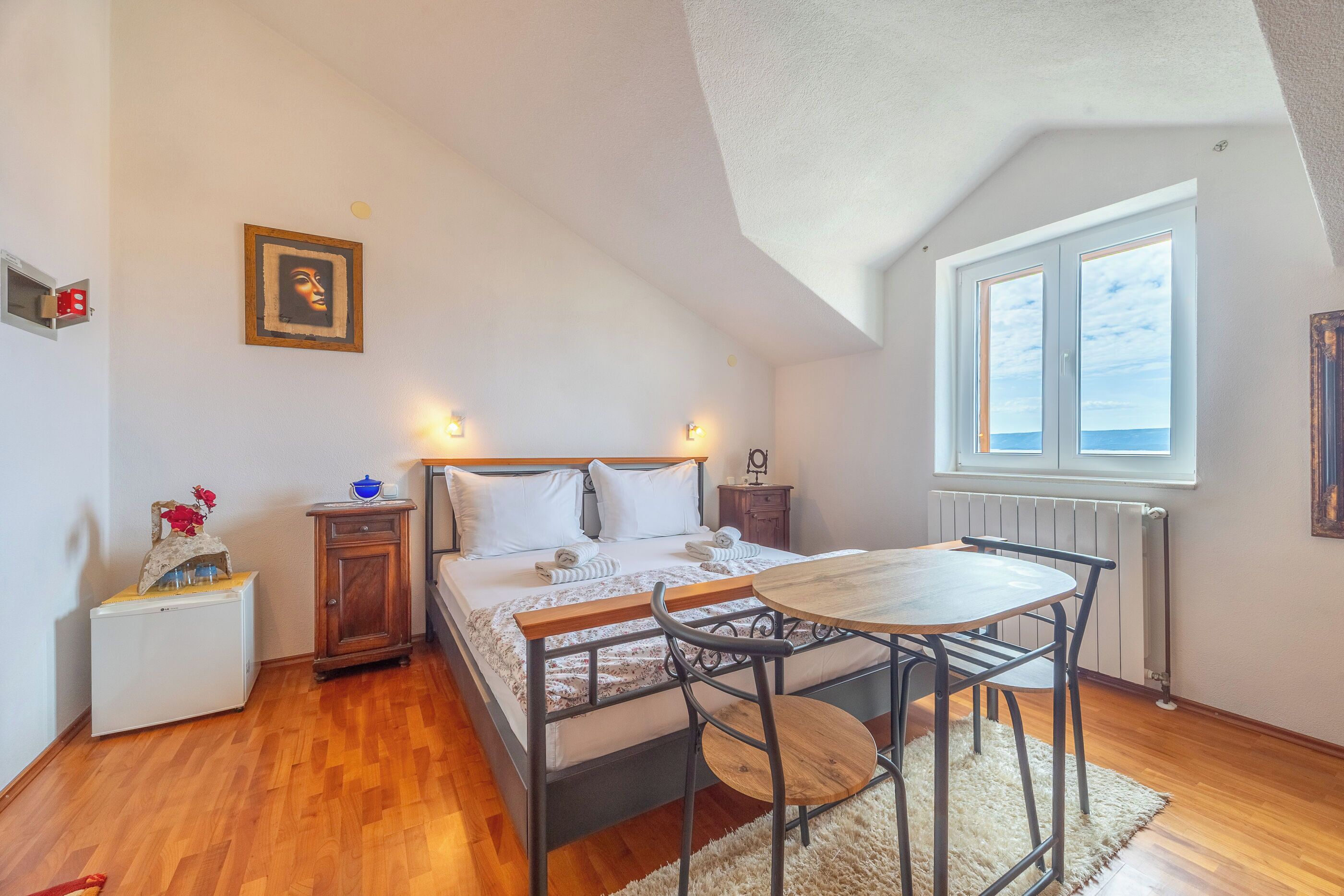 A bedroom with a bed, bedside tables, a small table with chairs, a radiator, and a window with a view.