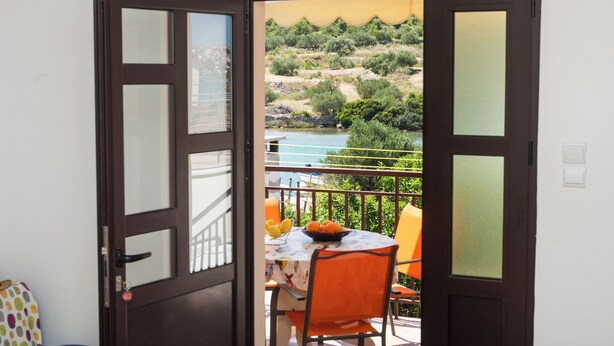 An open door to a balcony with a table set with oranges and lemons, overlooking a hillside and a body of water. - Apartments Hrvatic (Rogoznica)