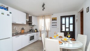 A modern kitchen with white cabinets, a built-in oven, and a dining area with a glass table and white chairs. - Apartments Hrvatic (Rogoznica)