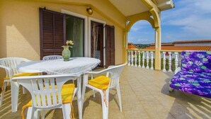 Un balcone con tavolo e sedie bianche, un divano con fiori viola e vista sugli edifici sullo sfondo.