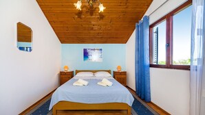 A bedroom with a wooden ceiling, a bed with a blue bedspread, two nightstands with lamps, a window with curtains, and a painting on the wall. - Apartment Sime (Nin)